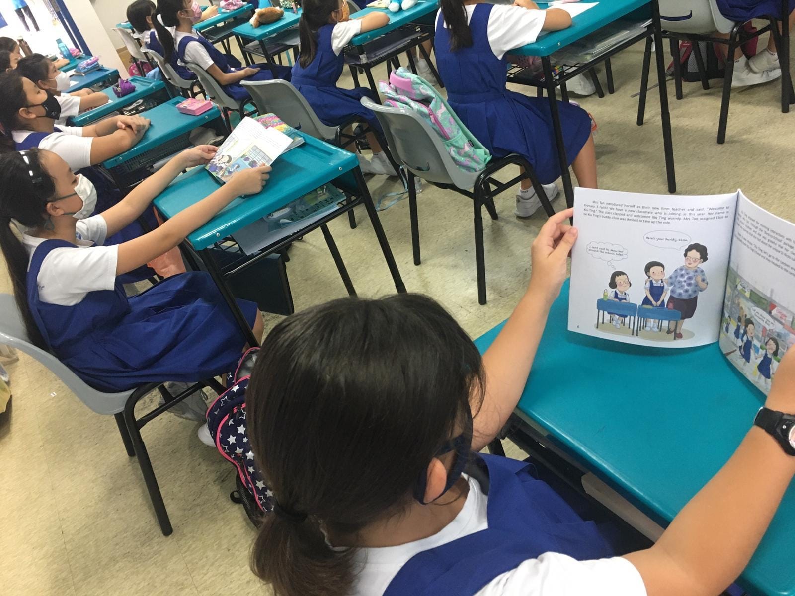 Girls in blue uniforms sit at teal desks in a classroom, reading illustrated storybooks.